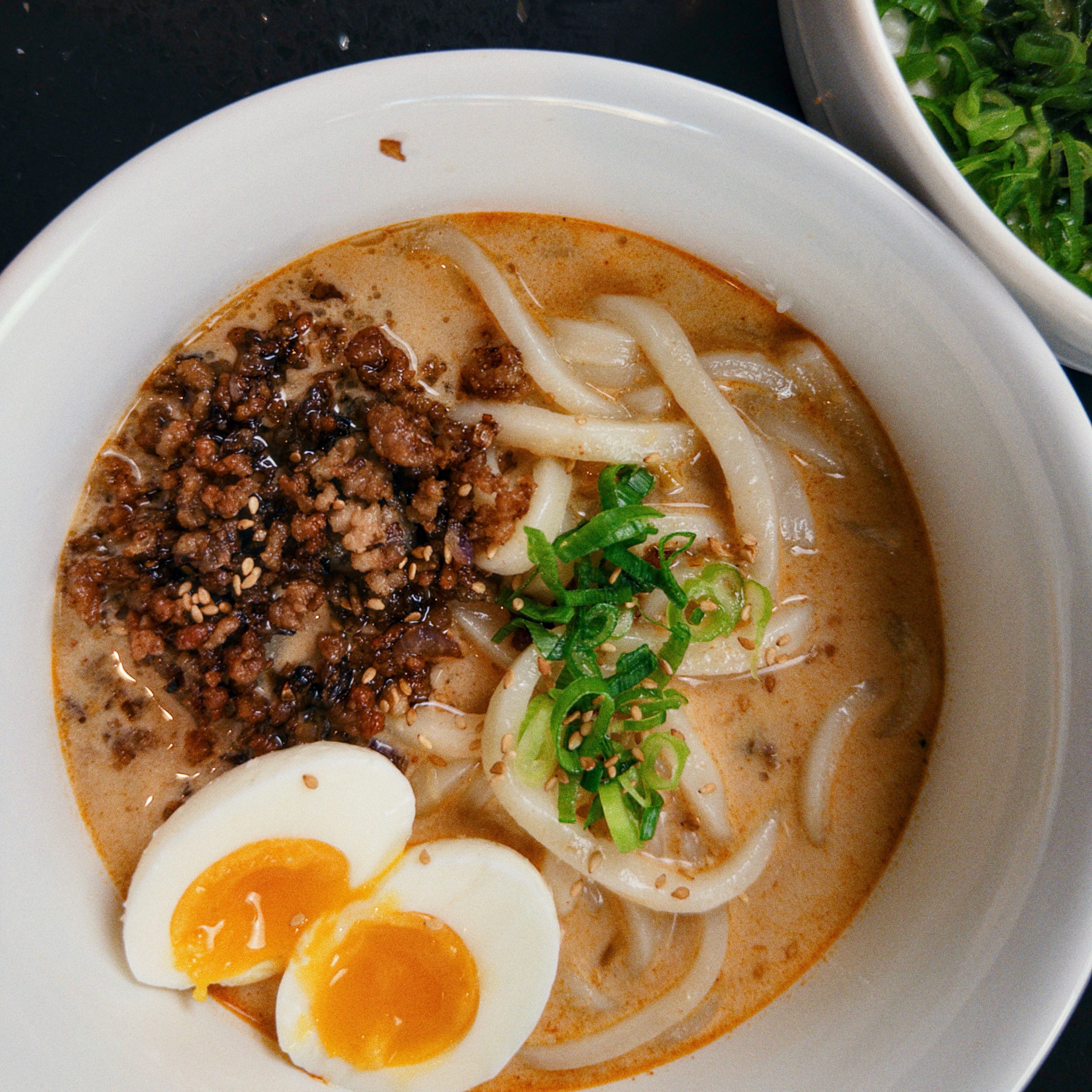 Ramen cremoso ao curry coberto com carne suína moída caramelizada, ovo com gema cremosa, cebolinha, gergelim e molho de pimenta